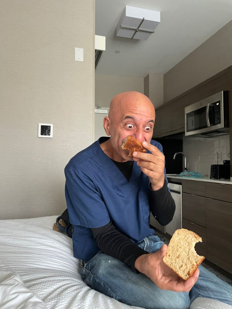 A man with a surprised expression sniffs bread while sitting on a bed